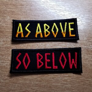May include: Two rectangular black embroidered patches. The top patch reads "AS ABOVE" in yellow and orange thread. The bottom patch reads "SO BELOW" in red thread. The patches are on a wooden surface.