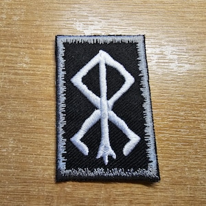 Pode incluir: Símbolo branco bordado em um remendo preto com borda cinza. O símbolo é uma runa estilizada.