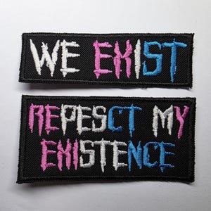 Op de afbeelding: Twee rechthoekige, zwart geborduurde patches met gestikte randen. De bovenste patch bevat de tekst WE EXIST in wit, roze en blauw. De onderste patch bevat de tekst REPESCT MY EXISTENCE in wit, roze en blauw in een grillig lettertype.