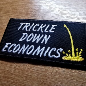 May include: Black embroidered patch with white text that reads "Trickle Down Economics". A yellow stylized illustration of a stream of liquid dripping down from the top of the patch.