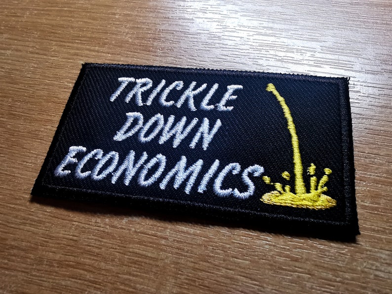 May include: Black embroidered patch with white text that reads "Trickle Down Economics". A yellow stylized illustration of a stream of liquid dripping down from the top of the patch.