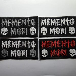 Può includere: Quattro toppe rettangolari ricamate su sfondo nero. Ogni toppa presenta la scritta MEMENTO MORI in un carattere stilizzato con un teschio sul lato sinistro e destro. Tre toppe hanno ricami bianchi, una ha scritte rosse con teschi bianchi.