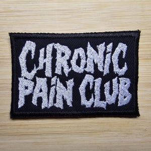 Op de afbeelding: Zwarte geborduurde patch met witte tekst die "Chronic Pain Club" luidt.