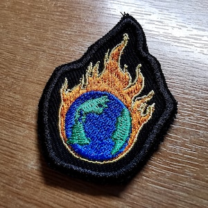 Op de afbeelding: Een zwarte geborduurde patch met een blauwe en groene wereld die omhuld is door oranje vlammen. De wereld is omgeven door een gele omtrek.