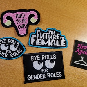 May include: Five embroidered iron-on patches with feminist themes. The patches are black, white, pink, and blue. The patches feature the text "Mind Your Own", "The Future is Female", "Eye Rolls for Gender Roles", "Eye Rolls for Gender Roles", and "Never Again".