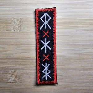 Puede incluir: Un parche bordado negro y rojo con runas blancas y cruces rojas. El parche es rectangular y tiene un borde rojo.