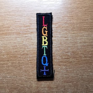 Könnte beinhalten: Ein schwarzer Stoff-Patch mit den Buchstaben LGBTQ+ in Regenbogenfarben gestickt.