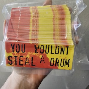 Op de afbeelding: Een stapel rechthoekige stickers in een doorzichtige plastic zak. De stickers hebben een rood-gele gradiënt ontwerp. De tekst "YOU WOULDNT STEAL A DRUM" is in zwart gedrukt op een rood-gele achtergrond.
