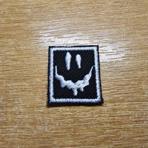 May include: A black and white embroidered patch with a white smiley face with three teeth. The patch is surrounded by a white border.