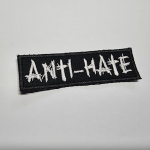 May include: A black rectangular patch embroidered with the words "ANTI-HATE" in white. The text is in a distressed, handwritten style. The patch is designed to be sewn or ironed onto clothing or accessories.