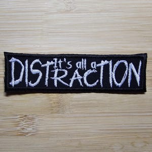 Puede incluir: Parche bordado en blanco y negro con el texto "It's all a distraction" en letras blancas sobre fondo negro.