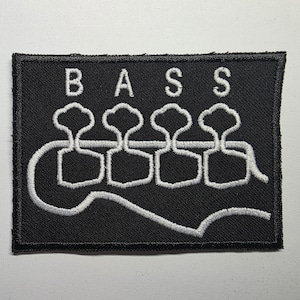 May include: Black rectangular patch featuring the word "BASS" in white capital letters. Below the letters are four white stylized bass guitar tuning pegs and a white bass guitar string.