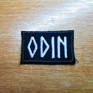 Puede incluir: Un parche rectangular negro con la palabra "ODIN" bordada en blanco. Las letras están en una fuente estilizada, con la "O" con forma de diamante. El parche es probablemente para ropa o accesorios.