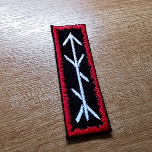 May include: A black embroidered patch with a white symbol of an arrow pointing upwards. The patch has a red border.