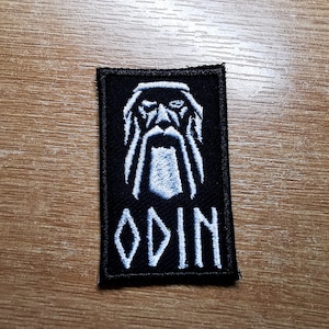 Könnte beinhalten: Ein schwarzer gestickter Aufnäher mit einer weißen Umrandung von Odins Gesicht und dem Wort "ODIN" darunter.