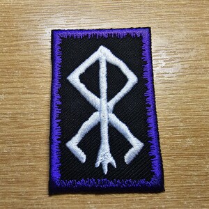 May include: White embroidered symbol on a black patch with a purple border. The symbol is a stylized rune.