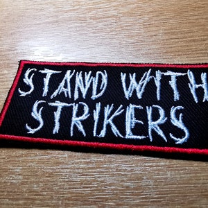 May include: Black embroidered patch with red stitching around the edges. The patch says "Stand with strikers" in white lettering.