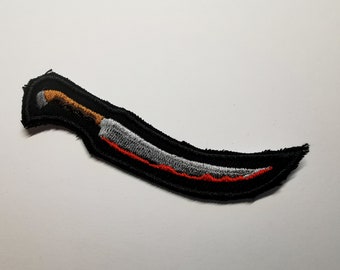 May include: A black embroidered patch of a curved knife with a brown handle, a silver blade, and a red blood stain.
