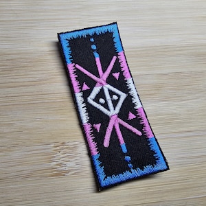 Op de afbeelding: Een zwarte geborduurde patch met een wit symbool en roze en blauwe randen. Het symbool is een gestileerd geometrisch ontwerp met een centrale ruitvorm.