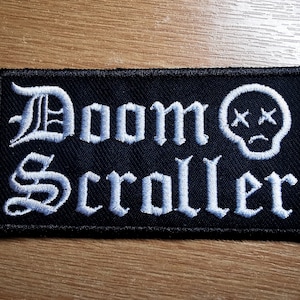 May include: White embroidered patch on a black background with the text "Doom Scroller" in a gothic font and a white skull with crossbones and a sad face.