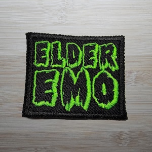 Może przedstawiać: Zielona haftowana naszywka z napisem "Elder Emo" na czarnym tle.