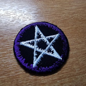Puede incluir: Un parche circular negro con un borde morado en el que se borda un pentagrama blanco.