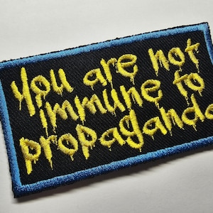 Può includere: Toppa ricamata con la scritta "You are not immune to propaganda" in giallo gocciolante su sfondo nero. La toppa ha un bordo blu.