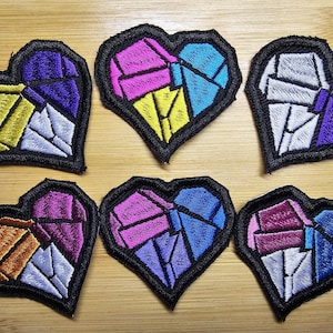 以下が含まれることがあります： ステンドグラスのデザインが施された、ハート型の刺繍パッチが6つ。各パッチは黒い縁取りと、紫、青、ピンク、黄色、オレンジ、白の色合いのカラフルな幾何学模様が特徴です。パッチは木製の表面に配置されています。