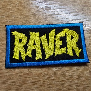 May include: A black and yellow embroidered patch with the word "RAVER" in a bold, stylized font. The patch has a blue border.