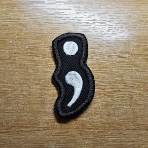Op de afbeelding: Zwarte stoffen patch met een wit geborduurd puntkomma symbool.