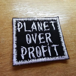 Puede incluir: Parche bordado negro con costuras blancas. El parche dice "Planet Over Profit".