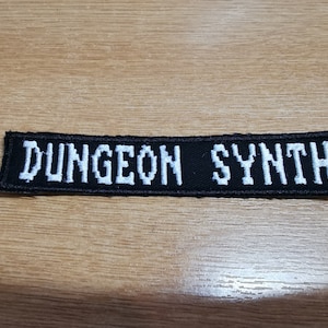 May include: Black embroidered patch with the words "DUNGEON SYNTH" in white block letters. The patch is rectangular and has a black border. The patch is on a light brown wooden surface.
