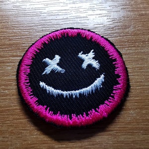 May include: A black embroidered patch with a white smiley face with X's for eyes. The patch has a pink border.