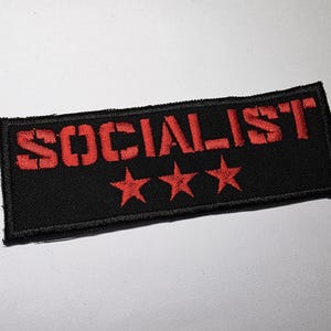 May include: Black rectangular patch featuring the word "SOCIALIST" in red, block letters, and three red stars. This embroidered patch could be used to customize clothing or accessories.
