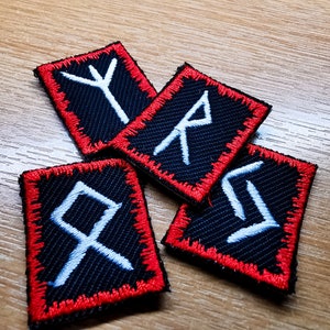 Könnte beinhalten: Vier schwarze Stoffpatches mit roten Nähten an den Rändern. Jeder Patch zeigt ein weiß gesticktes Runensymbol.