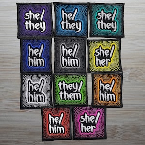 May include: Nine embroidered iron-on patches with various pronoun combinations, including "she/they", "he/him", "they/them", and "she/her". The patches are on a wooden surface.