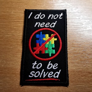 Könnte beinhalten: Schwarzer gestickter Aufnäher mit weißem Text, der "Ich brauche keine Lösung" lautet. Ein roter Kreis mit einem diagonalen roten Strich enthält ein buntes Puzzle-Motiv.