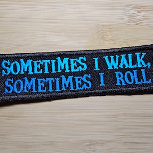 Puede incluir: Parche rectangular negro bordado con el texto "SOMETIMES I WALK, SOMETIMES I ROLL" en letras degradadas azules. El parche est&aacute; sobre una superficie de madera marr&oacute;n claro.