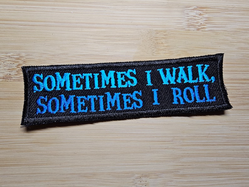 Puede incluir: Parche rectangular negro bordado con el texto "SOMETIMES I WALK, SOMETIMES I ROLL" en letras degradadas azules. El parche est&aacute; sobre una superficie de madera marr&oacute;n claro.