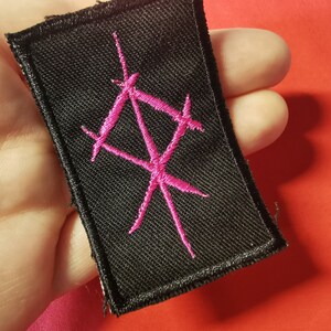 May include: A black fabric patch with a pink embroidered symbol. The symbol is a stylized design of intersecting lines.