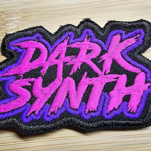 Pode incluir: Patch bordado com as palavras "DARK SYNTH" em rosa vibrante, contornado em roxo, sobre um fundo preto. O patch tem uma borda irregular e é para roupas ou acessórios.