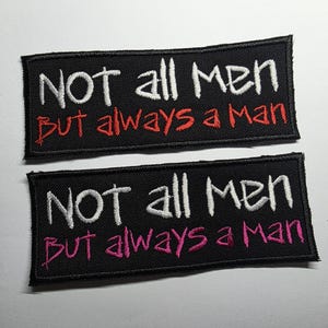 Puede incluir: Dos parches rectangulares negros con texto bordado en blanco que dice "NOT all MEN" encima de "But always a man" en rojo y rosa. Los parches tienen un borde cosido.