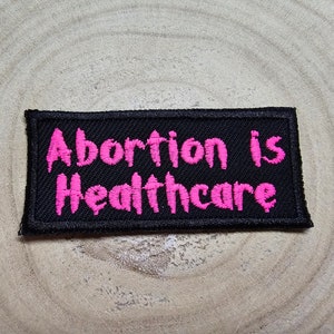 May include: Black fabric patch with pink embroidered text that reads "Abortion is Healthcare".