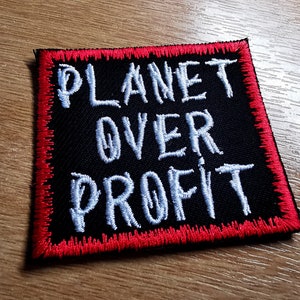 Puede incluir: Un parche negro con las palabras "PLANET OVER PROFIT" bordadas en blanco. El parche tiene un borde rojo y está diseñado para coserlo en ropa o accesorios. El parche es cuadrado con esquinas redondeadas.