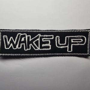 Op de afbeelding: Zwart rechthoekig embleem met de woorden "WAKE UP" in wit geborduurd. De patch heeft een wit gestikte rand en een zwarte achtergrond. Geschikt voor kleding of accessoires.