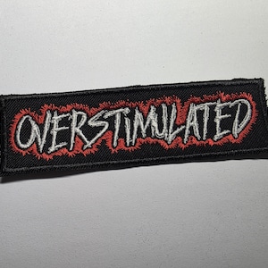 May include: Black rectangular patch with the word "OVERSTIMULATED" embroidered in white thread. The text is outlined with red stitching, creating a bold contrast. The patch is likely designed to be sewn or ironed onto clothing or accessories.