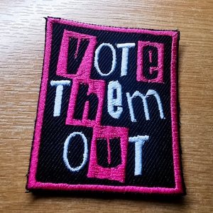 May include: A black fabric patch with pink and white embroidery. The patch says "Vote Them Out" in a bold, blocky font.