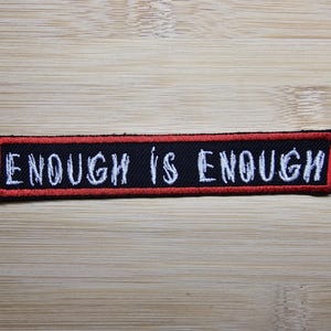 以下が含まれることがあります： 白文字で「Enough is Enough」と書かれた黒と赤の刺繍パッチ