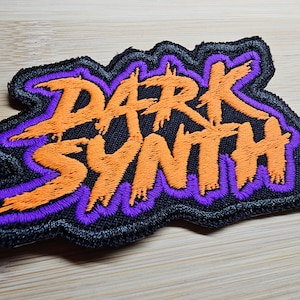 Puede incluir: Un parche bordado negro con las palabras "DARK SYNTH" en letras naranja y morado.