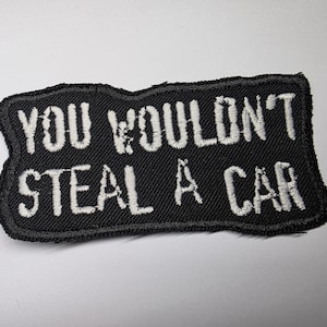 Op de afbeelding: Zwart rechthoekig embleem met witte geborduurde tekst: "YOU WOULDN'T STEAL A CAR". De patch heeft een zwarte rand en is ontworpen om op kleding of accessoires te worden genaaid.